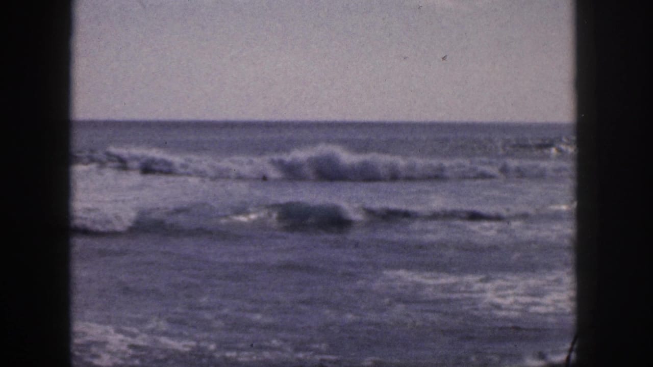 Ocean waves breaking under a pale sky, seen through dark vertical frame borders.
