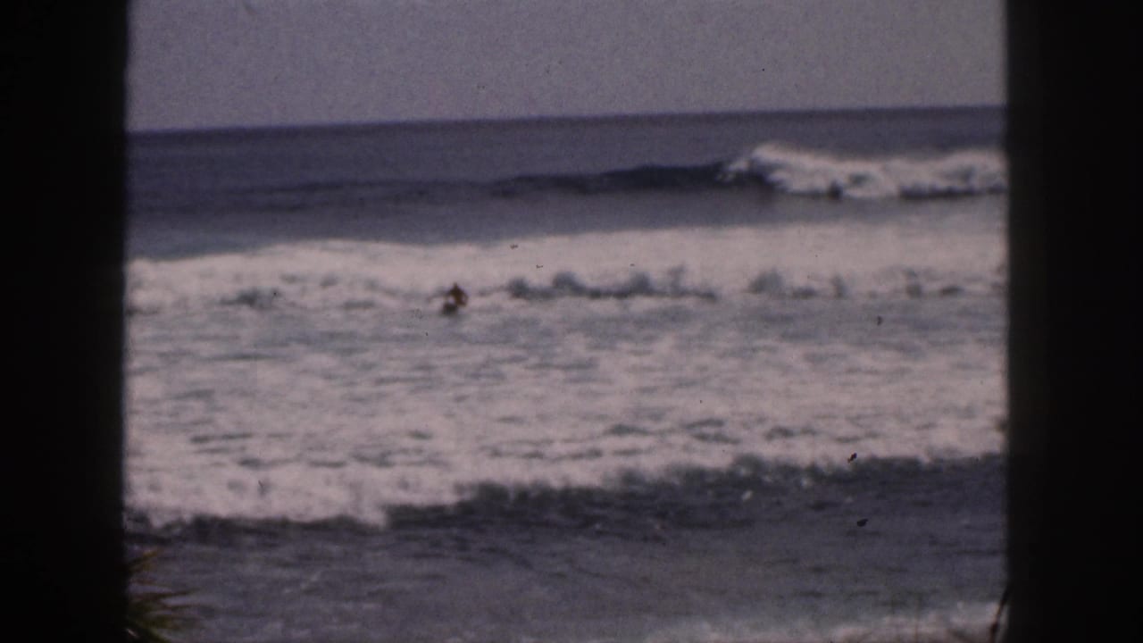 Distant prone surfer in breaking ocean waves seen through dark vertical framing.