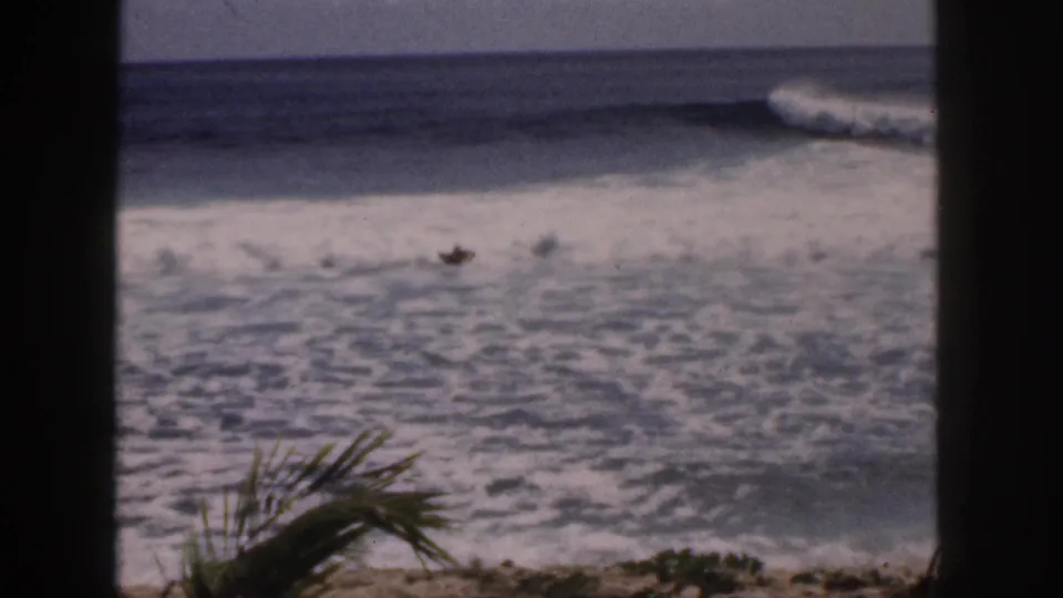 1959 Hawaii 8mm still of a prone surfer in whitewater, with palm fronds and dark window-like framing in the foreground.