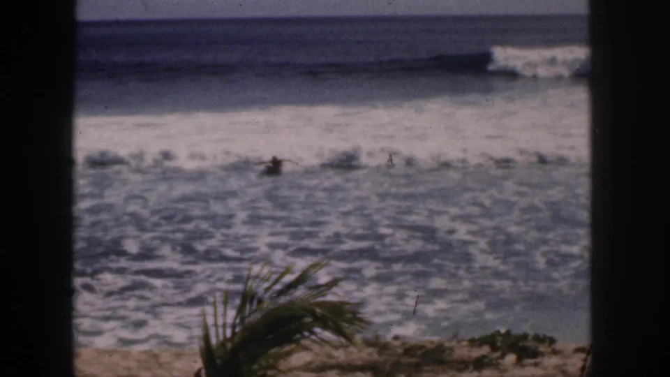 1959 Hawaii home-movie frame showing a small prone surfer near breaking waves, partly framed by palm fronds and dark vertical borders.