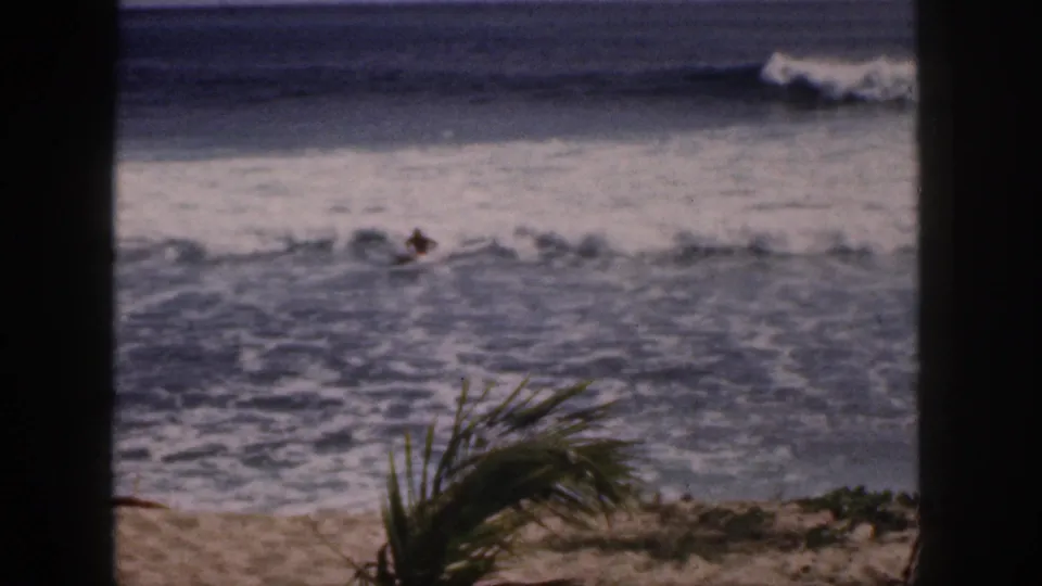 1959 Hawaii 8mm frame showing a distant beach rider or swimmer in breaking surf, viewed past dark framing and palm fronds.