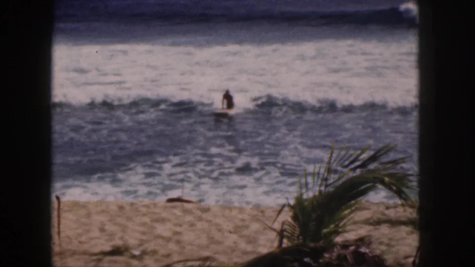 1959 Hawaii beach frame with a distant board rider upright in the surf, seen through a dark window or doorway-like foreground.