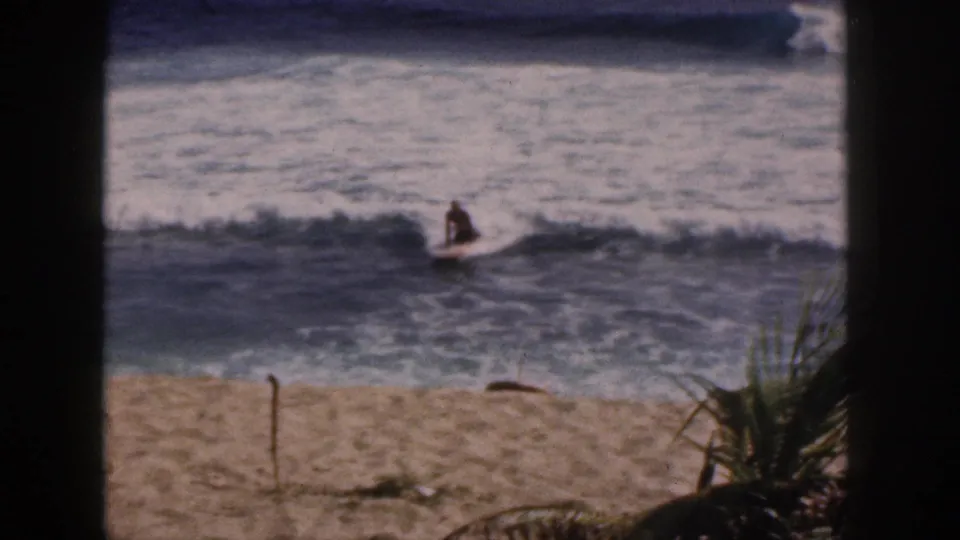1959 Hawaii 8mm surf scene showing a distant person on a board moving across a wave near palm fronds and sand.