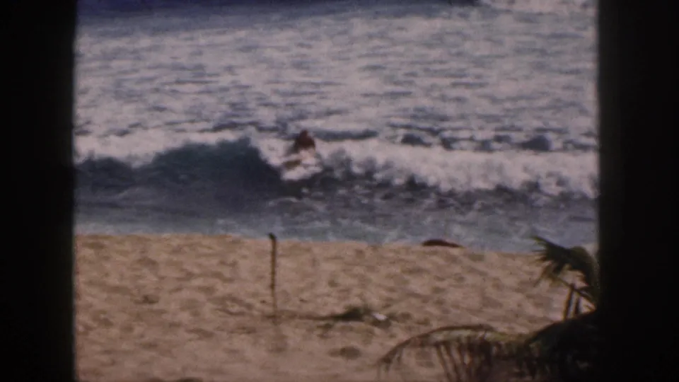 1959 Hawaii home-movie frame with a rider in foamy surf, framed by dark foreground edges and tropical beach plants.