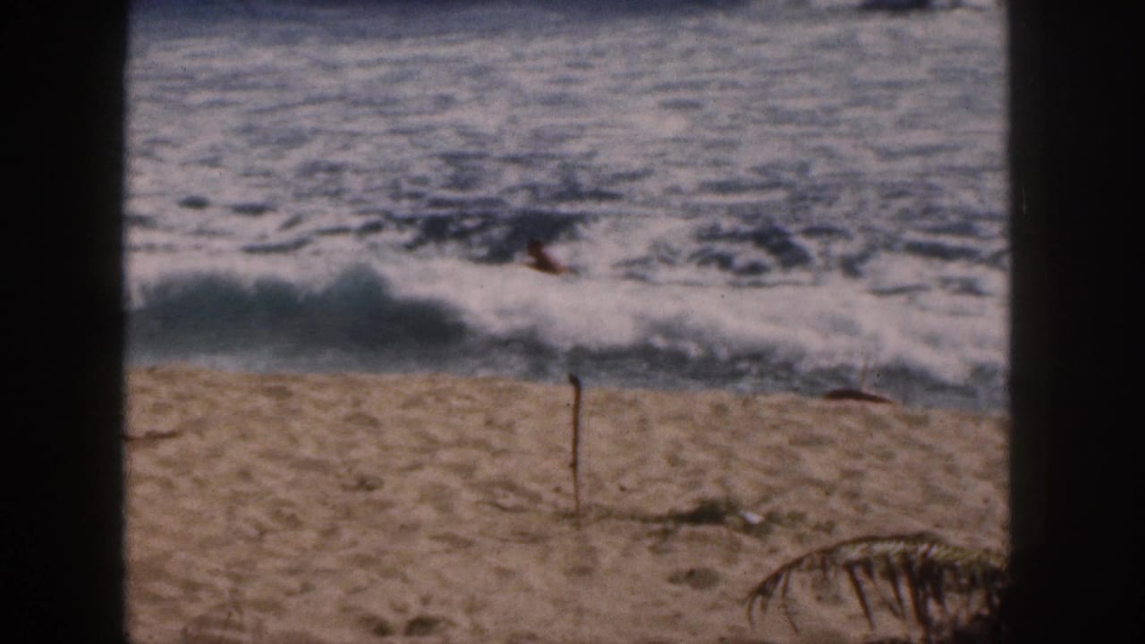 1959 Hawaii 8mm frame showing surf breaking near shore, a sandy beach foreground, and a small distant swimmer in the waves.