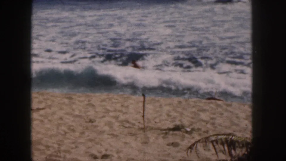 1959 Hawaii 8mm frame showing surf breaking near shore, a sandy beach foreground, and a small distant swimmer in the waves.