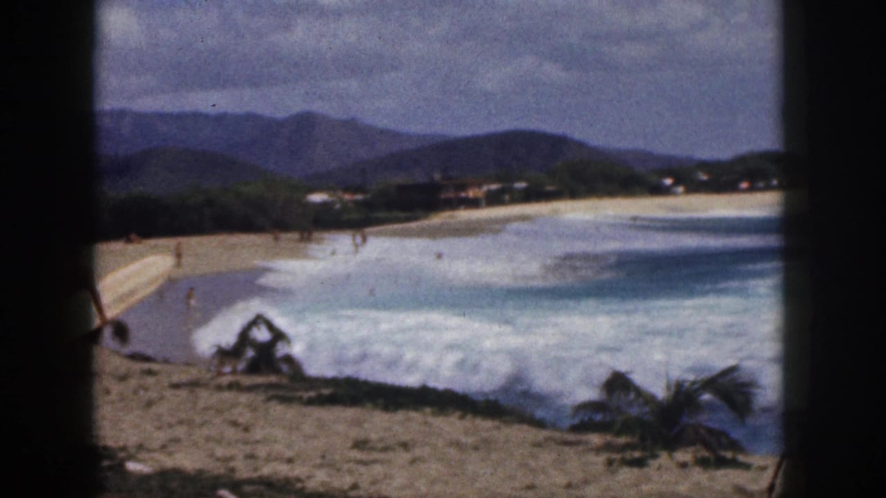 1959 Hawaii 8mm frame showing surf breaking along a sandy beach with mountains, palms, distant buildings, and small beachgoers.