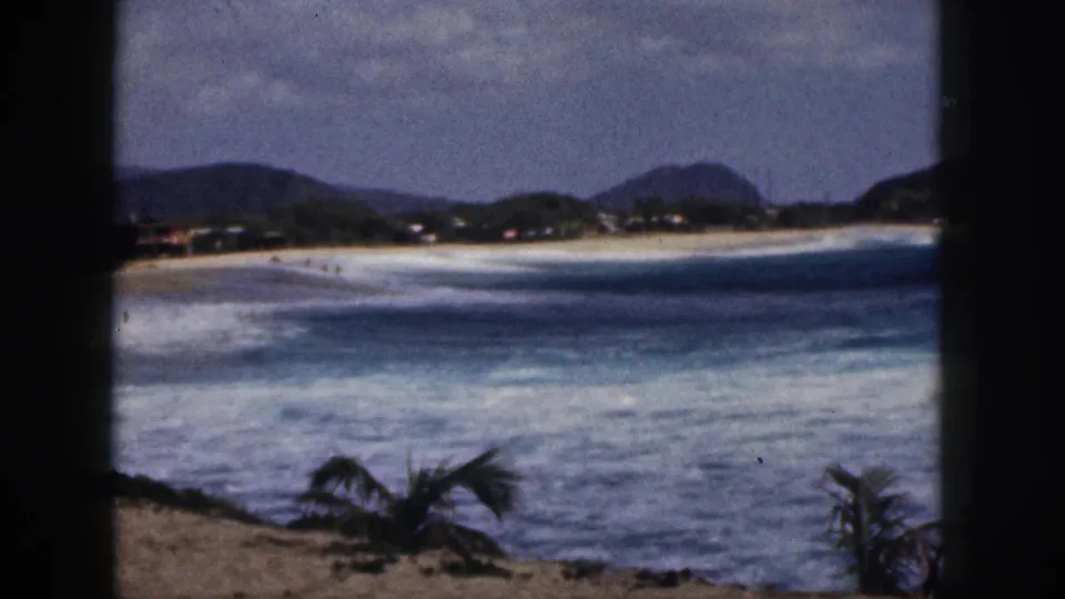 1959 Hawaii 8mm frame showing a broad beach and foamy surf from a high visitor viewpoint, with mountains and palms in view.