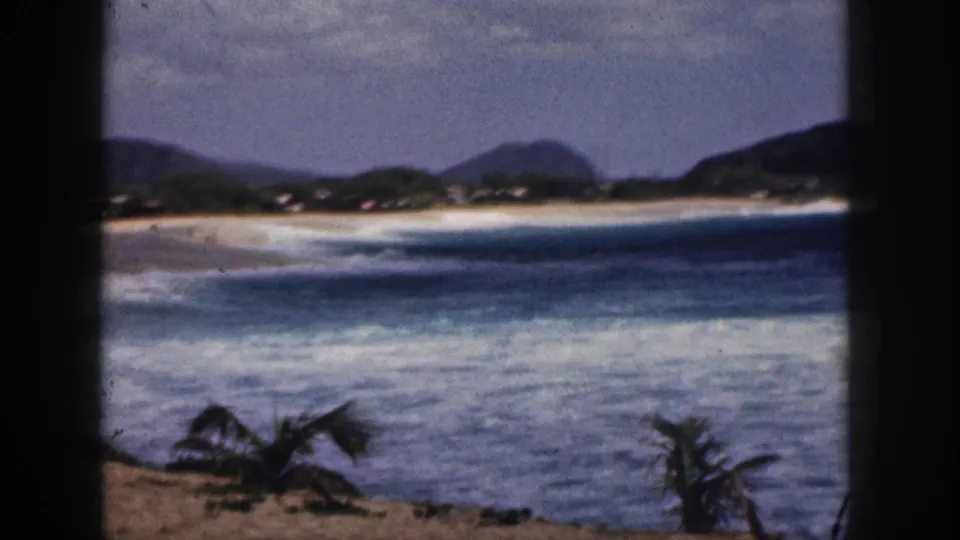 1959 Hawaii 8mm frame of rolling surf beside a sandy beach, with palms in the foreground and mountains behind the shoreline.
