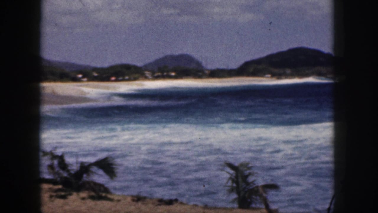 Blue ocean surf breaking near a sandy beach with distant green hills and dark frame edges.