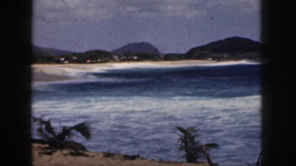 1959 Hawaii 8mm frame showing blue surf, a sandy shoreline, distant green hills, palm fronds, and dark vertical edges suggesting filming from behind a window or doorway.