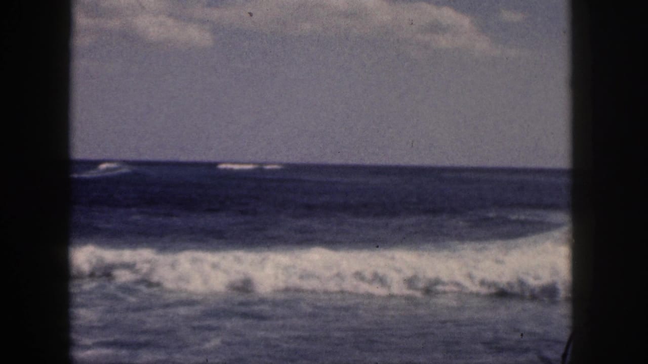 1959 Hawaii 8mm frame of a low line of breaking surf stretching across the ocean, with cloudy sky and dark frame borders.