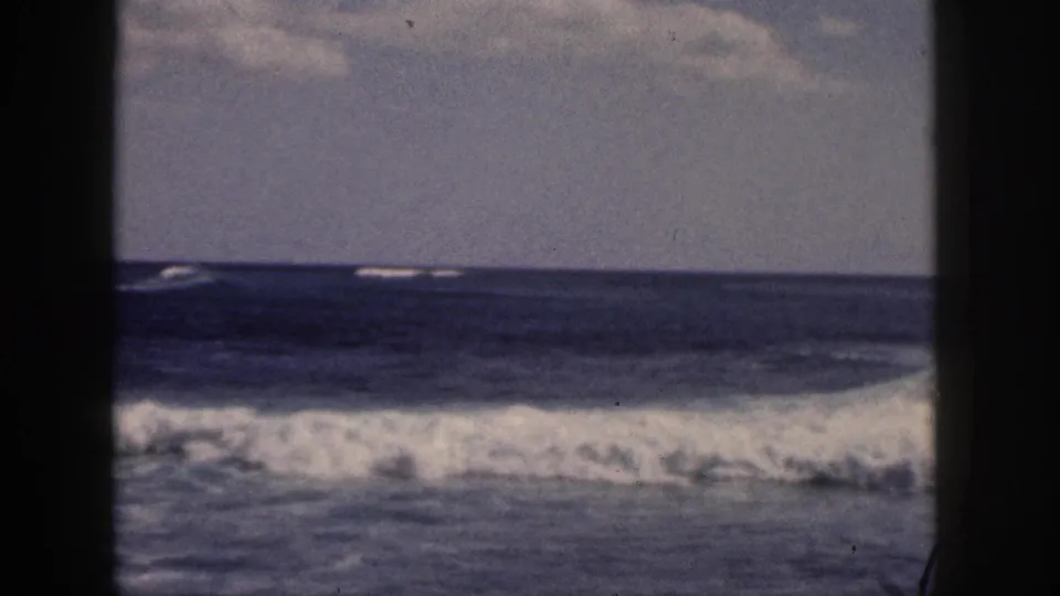 1959 Hawaii 8mm frame of a low line of breaking surf stretching across the ocean, with cloudy sky and dark frame borders.