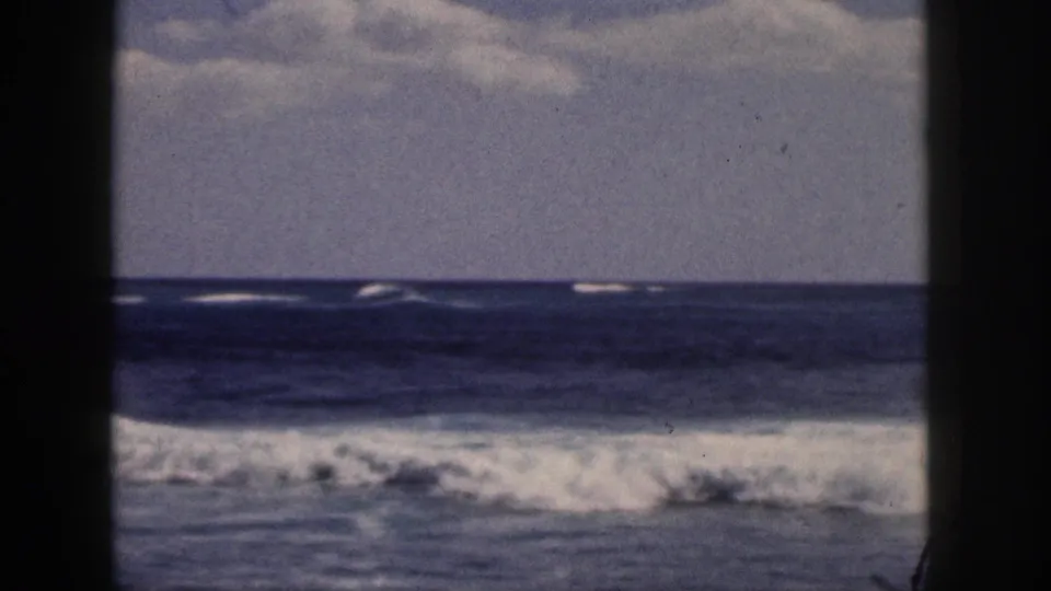 1959 Hawaii 8mm frame showing rolling ocean surf through dark vertical framing beneath a clouded sky.