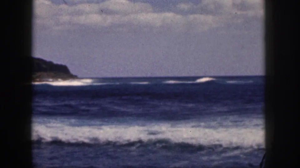 1959 Hawaii shoreline frame with a rocky headland at left, dark foreground framing, and white surf across blue water.