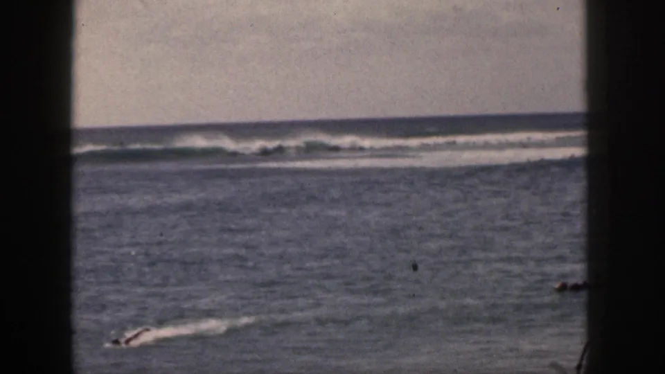 1959 Hawaii home-movie frame with a distant paddler or swimmer on open water, reef-like surf breaks, and dark vertical film borders.