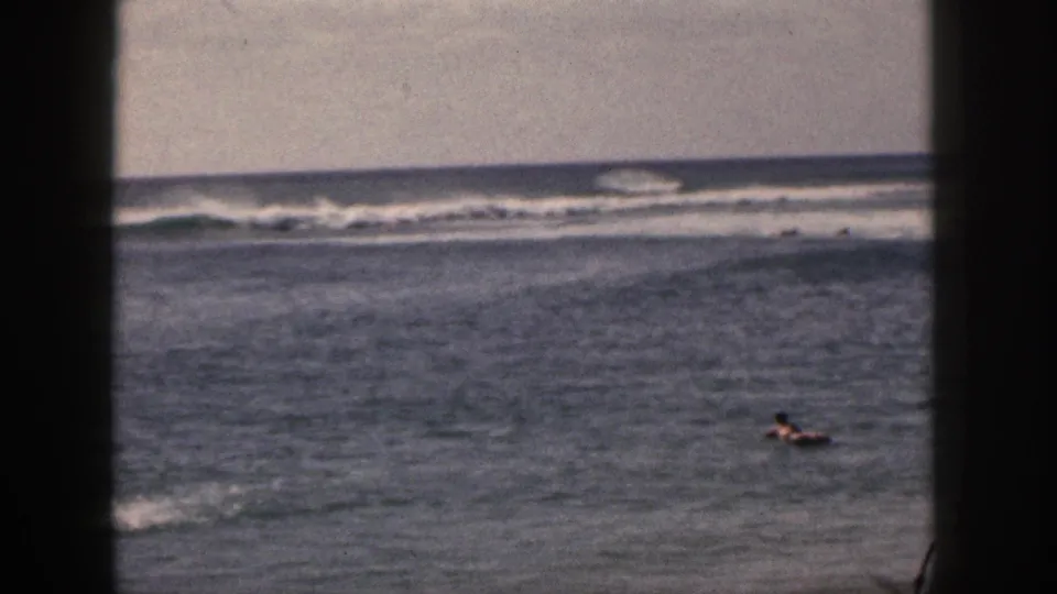 1959 Hawaii 8mm view of a distant person in the ocean, rolling waves, shoreline foam, muted color, and a dark film-frame divider.
