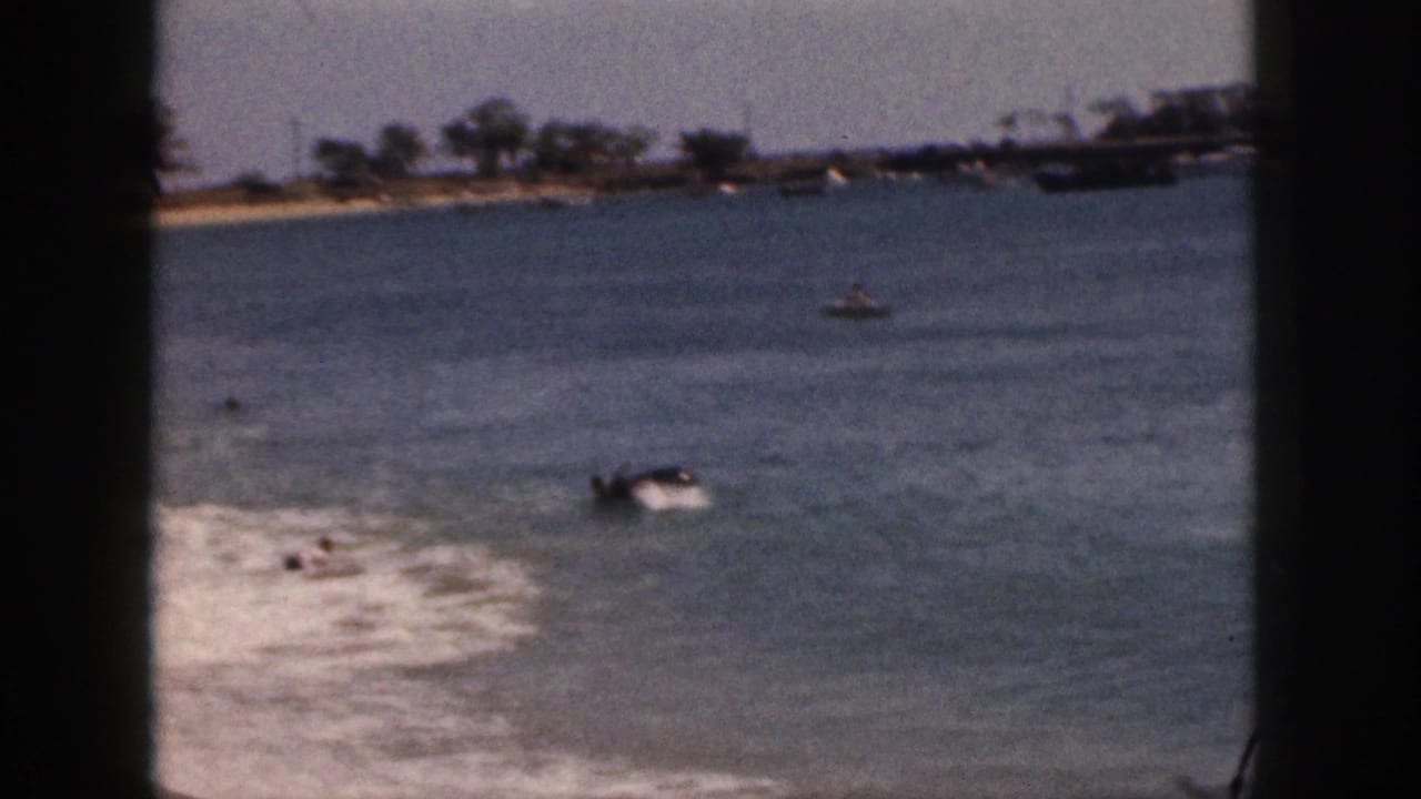 1959 Hawaii 8mm frame showing swimmers in shallow ocean water, a sandy edge, distant trees, and boats viewed through dark window bars.