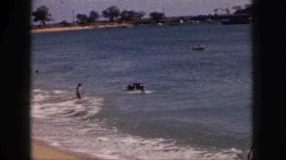 1959 Hawaii beach footage frame with a person wading in shallow surf, an inner tube nearby, and boats along the distant shoreline.