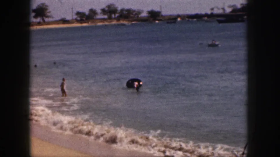 1959 Hawaii 8mm still with a swimmer near shallow surf, a dark inner tube, calm ocean water, and a tree-lined shoreline in the background.