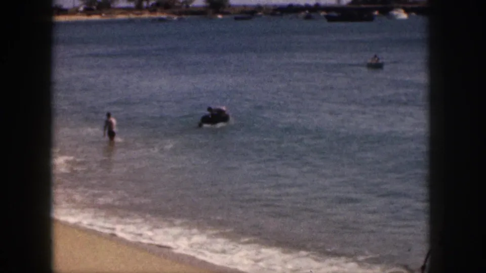 1959 Hawaii beach frame showing a person in shallow water, an inner tube to the side, low surf, and distant boats near the shoreline.