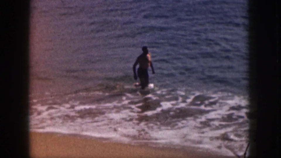 1959 Hawaii home-movie frame showing a man in dark swim trunks wading in shallow surf beside a sandy shoreline.