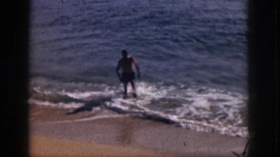 1959 Hawaii beach footage frame with a distant man in dark swim trunks moving through white surf toward the sand.