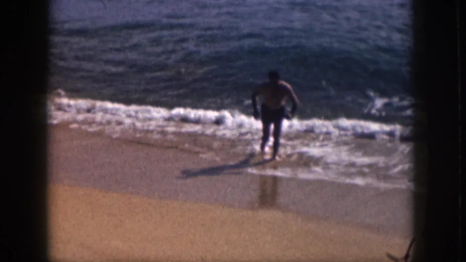 1959 Hawaii home-movie frame showing a man leaving the surf for the wet beach, with his shadow visible on the sand.
