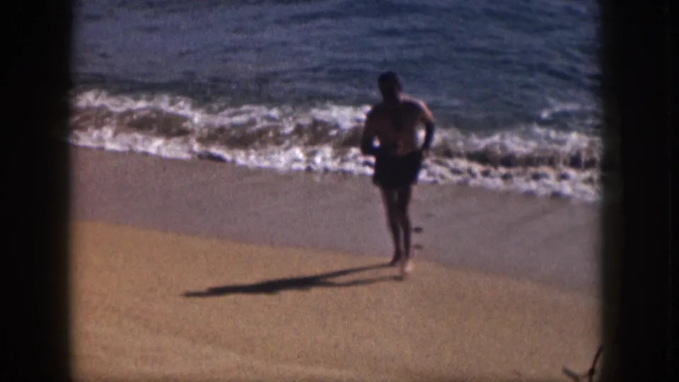 1959 Hawaii shoreline frame showing a man in dark swim trunks moving along the sunlit sand beside breaking surf.