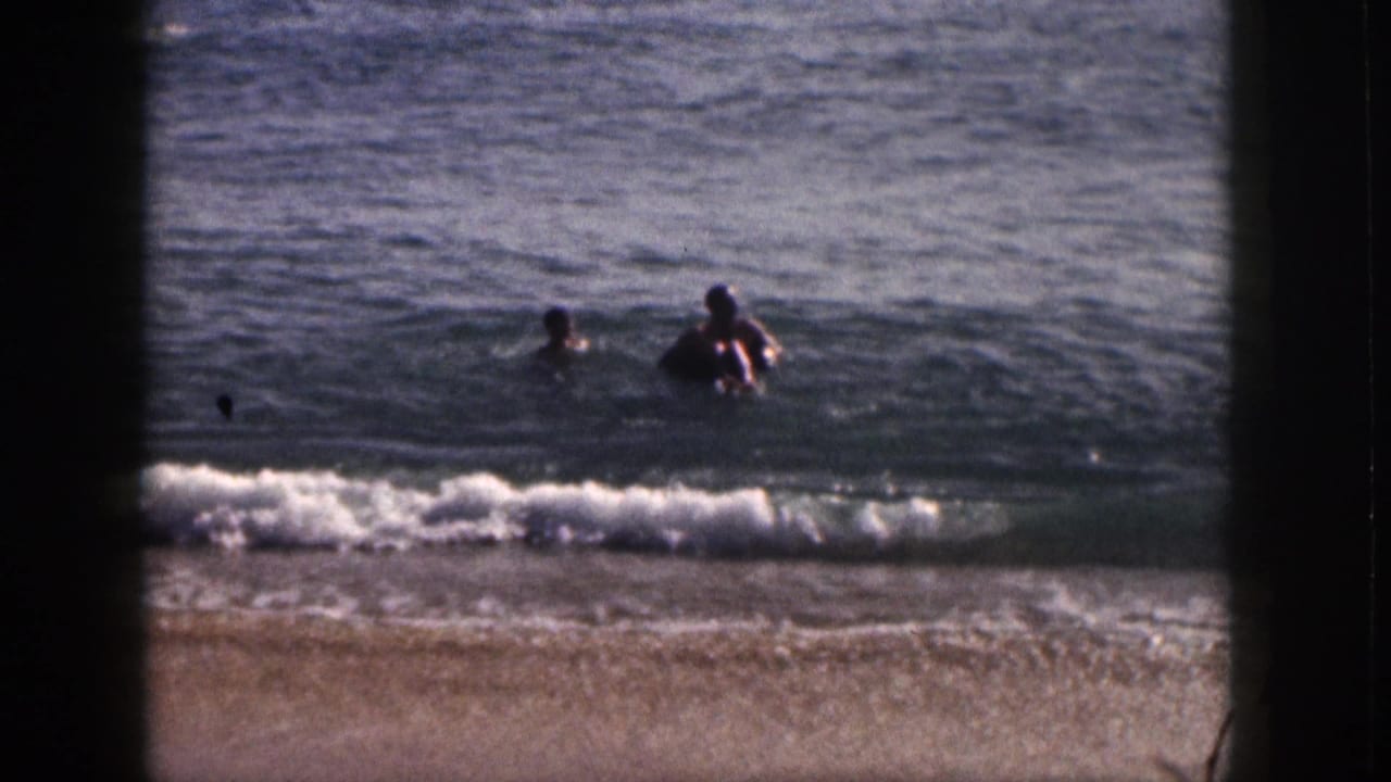 Two people swim near shore in 1959 Hawaii beach home-movie footage, framed by dark 8mm film borders.