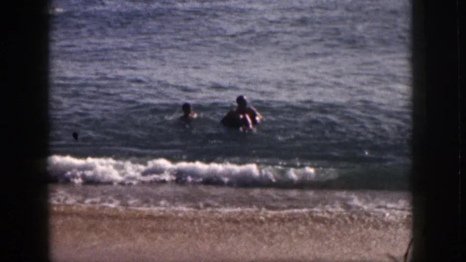 Two people swim near shore in 1959 Hawaii beach home-movie footage, framed by dark 8mm film borders.
