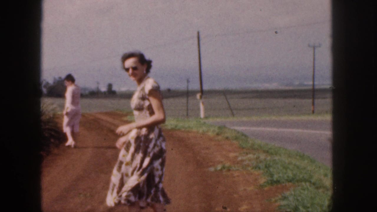 In 1959 Hawaii footage, two women in dresses walk on a red dirt road beside open fields and utility poles.