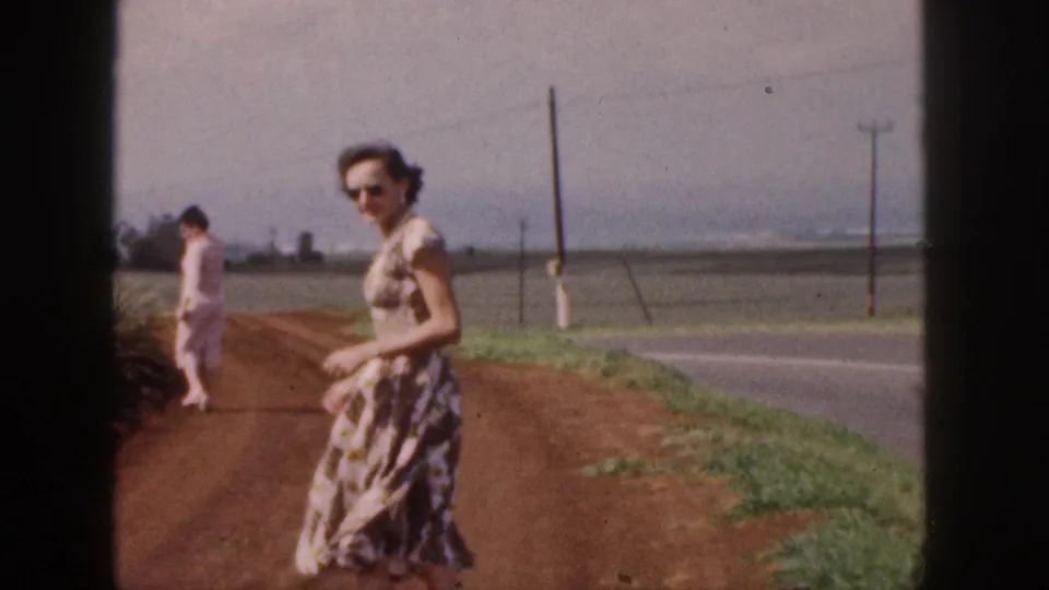 In 1959 Hawaii footage, two women in dresses walk on a red dirt road beside open fields and utility poles.