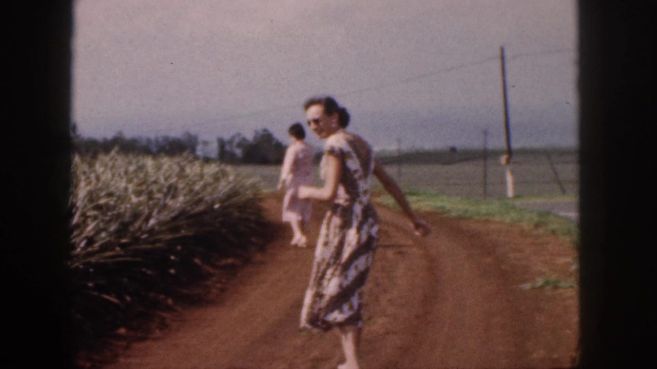 A woman turns toward the camera near pineapple rows while another walks ahead.