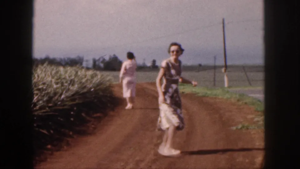 A woman in sunglasses walks toward the camera on a red dirt plantation road in Hawaii, with pineapple rows at left.