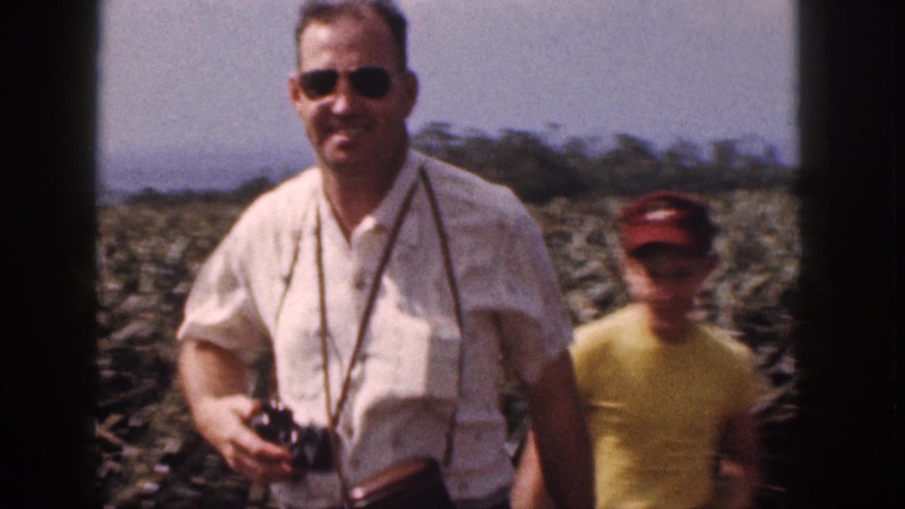Man wearing sunglasses and a camera stands in a pineapple field with another person behind him.