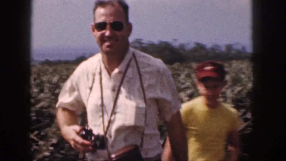 In 1959 Hawaii, a man in sunglasses with a camera around his neck stands before rows of pineapple plants.