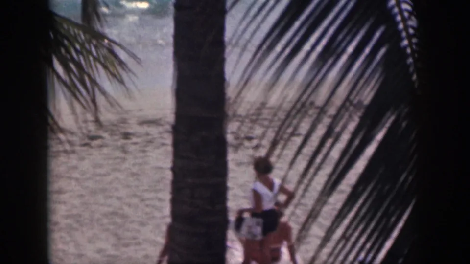 1959 Hawaii beach frame showing a woman in a white top and dark shorts walking near palm trunks and surf.