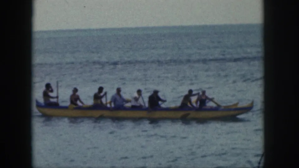 A home-movie view of the yellow outrigger canoe moving side-on, with the crew paddling over gently textured water.