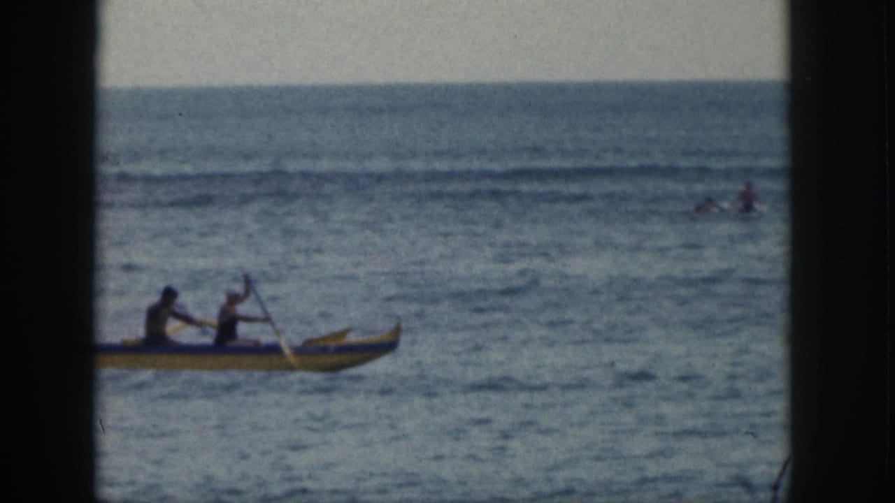 In 1959 Hawaii footage, two paddlers move a yellow outrigger canoe across calm ocean water with distant figures in the surf.