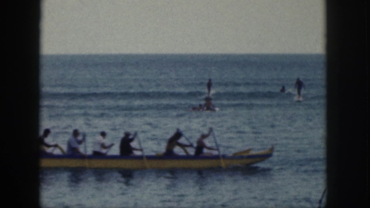 Several people ride in a long yellow outrigger canoe near the bottom of the frame, with other figures visible farther out in the surf.