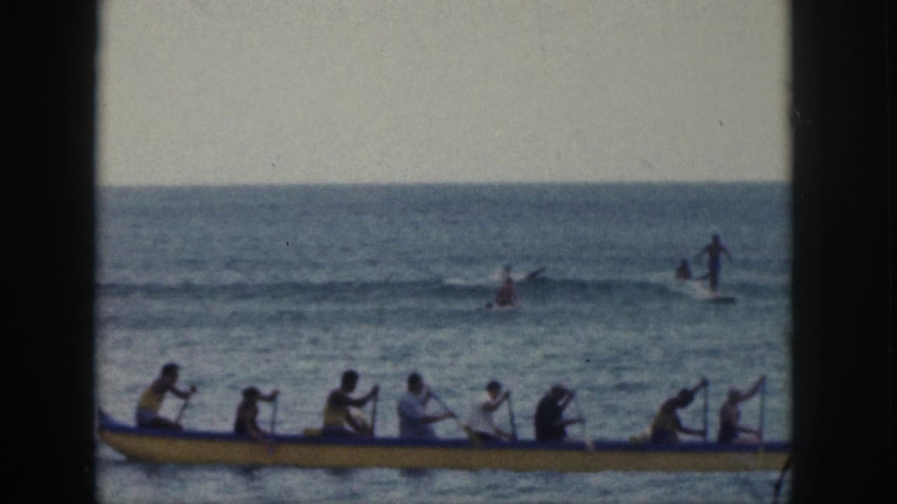 A long yellow outrigger canoe carrying multiple paddlers crosses the lower frame while distant surfers or swimmers appear beyond the small waves.