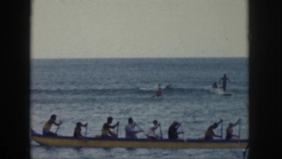 A long yellow outrigger canoe carrying multiple paddlers crosses the lower frame while distant surfers or swimmers appear beyond the small waves.