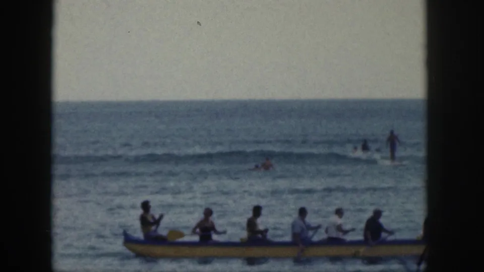 1959 Hawaii home-movie frame showing a yellow outrigger canoe with several paddlers on the ocean, with swimmers or surfers farther offshore.