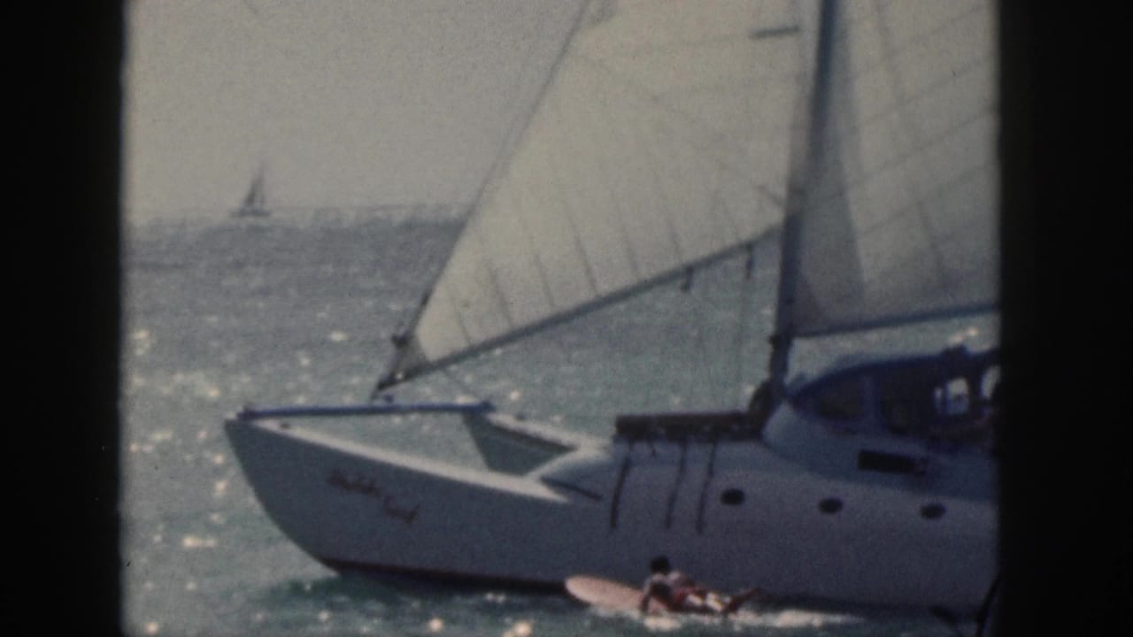 White sailboat with large sails on bright ocean water, with a surfer nearby.