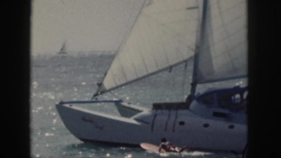 1959 Hawaii frame showing a white sailboat under large sails on sunlit water, with a person on a surfboard near the hull.