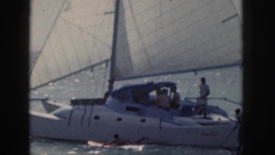 1959 Hawaii home-movie frame showing a white sailboat with people on deck and a surfboard crossing the foreground water.
