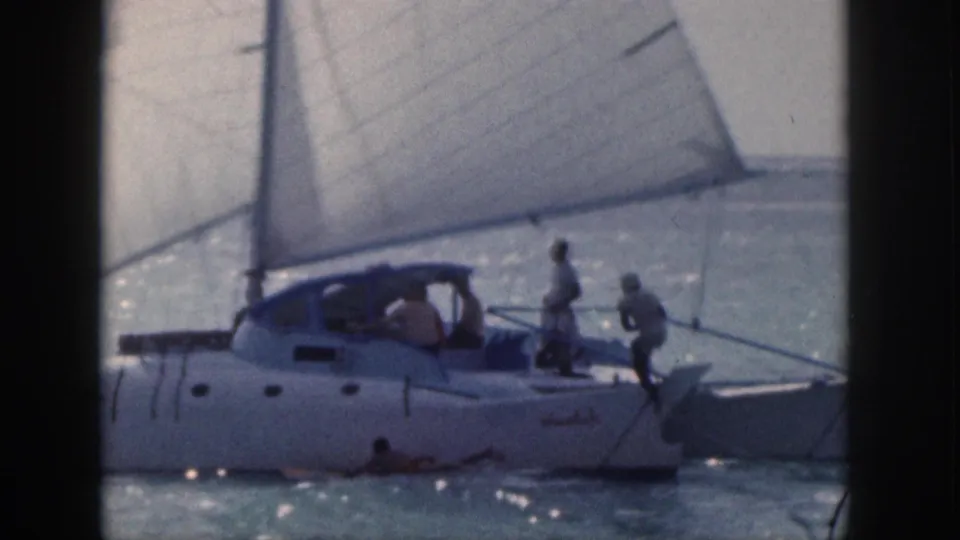 1959 Hawaii frame showing a white sailboat at close range, people on deck, and a swimmer or surfer in the water beside the hull.