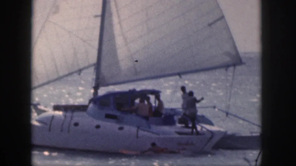 1959 Hawaii 8mm frame showing a white catamaran sailing on sunlit water with passengers aboard and a swimmer visible beside the hull.