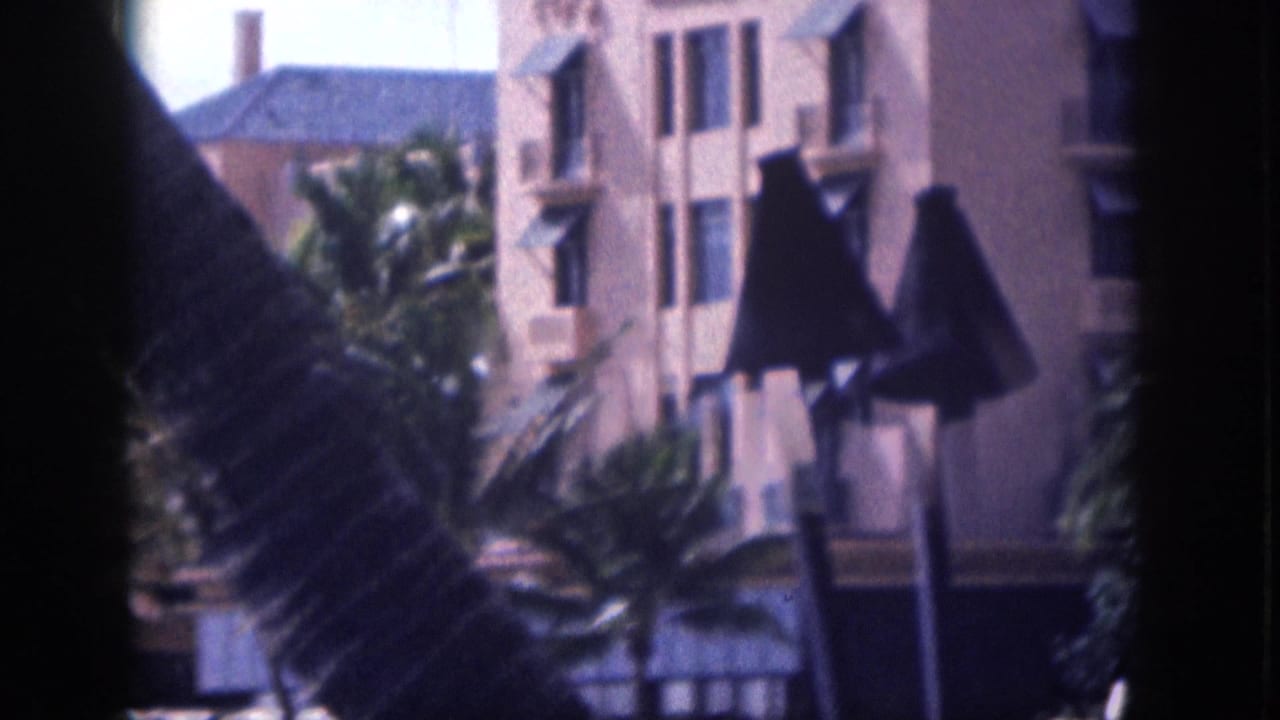 1959 Hawaii 8mm frame showing a pale Spanish Colonial Revival style building with awnings, palms, and dark foreground lamps.
